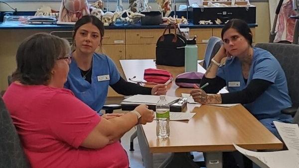 2 students in scrubs sitting at table interviewing mock patient